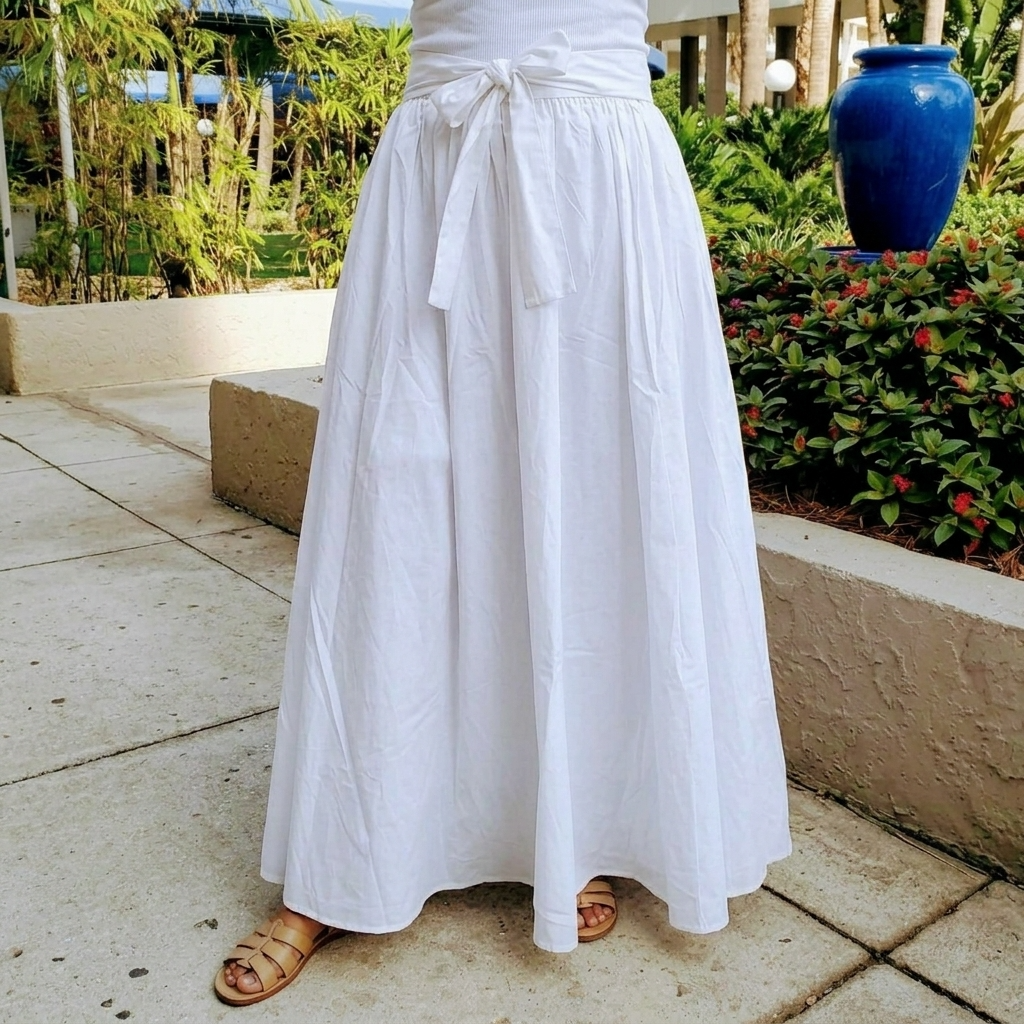 A person wearing a long white linen maxi skirt with a tie waist, paired with brown leather slide sandals, standing outdoors on a concrete patio with garden plants and a blue ceramic vase in the background.