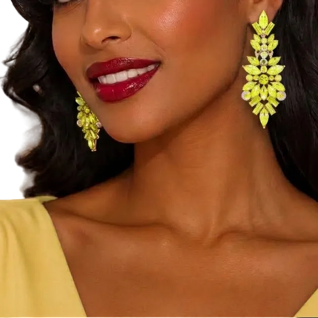A woman with styled dark hair and red lipstick wearing bright yellow floral drop earrings and a pale yellow top, photographed against a softly blurred indoor background with warm lights.