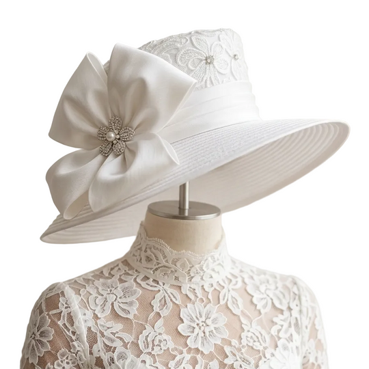 A white wide-brimmed sun hat with a large decorative bow and jeweled center, displayed on a cream lace dress form against a soft cream curtain background.