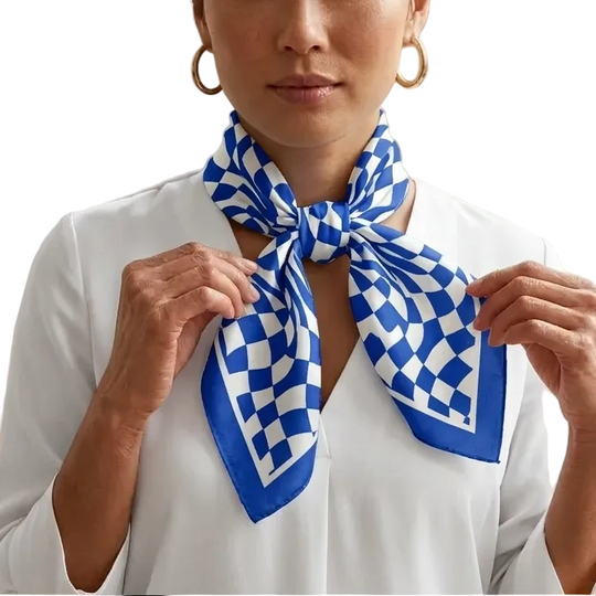 A woman wearing a white blouse and gold hoop earrings models a blue and white checkered silk scarf tied around her neck, set against a softly lit indoor background with a potted plant.