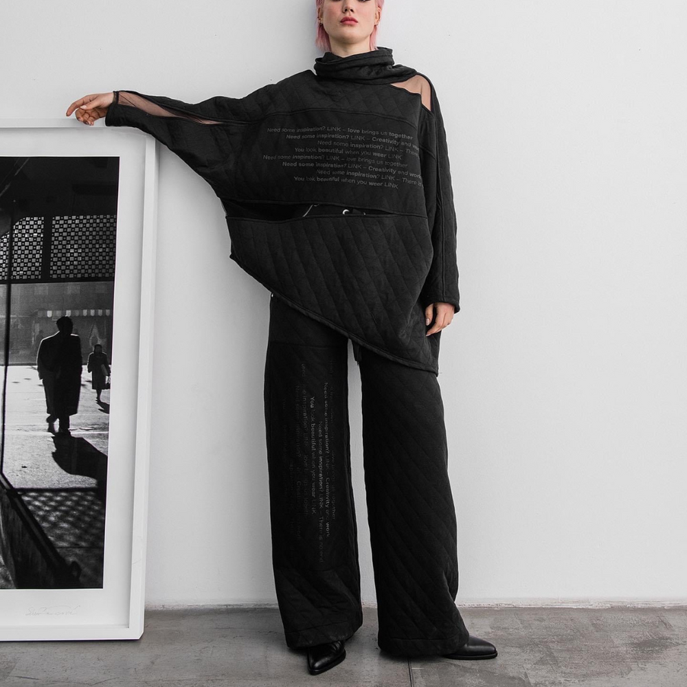 A woman modeling a black quilted top with matching trousers, standing in front of a photo frame. The top features text on the front.